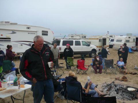 1988 Ford Bronco - People are gathered on a sandy beach with RVs and a large white SUV under an overcast sky.