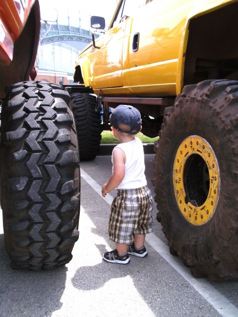 1994 Ford F-250 - A child stands between the enormous tires of a yellow monster truck, dwarfed by its size.