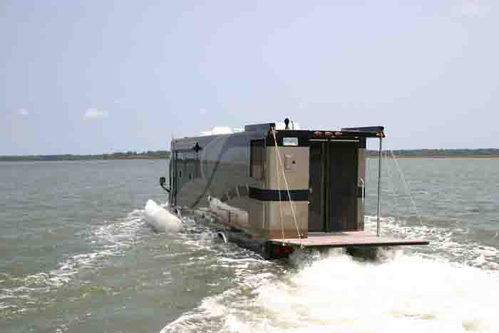 RV converted into a houseboat moving through water, rear view