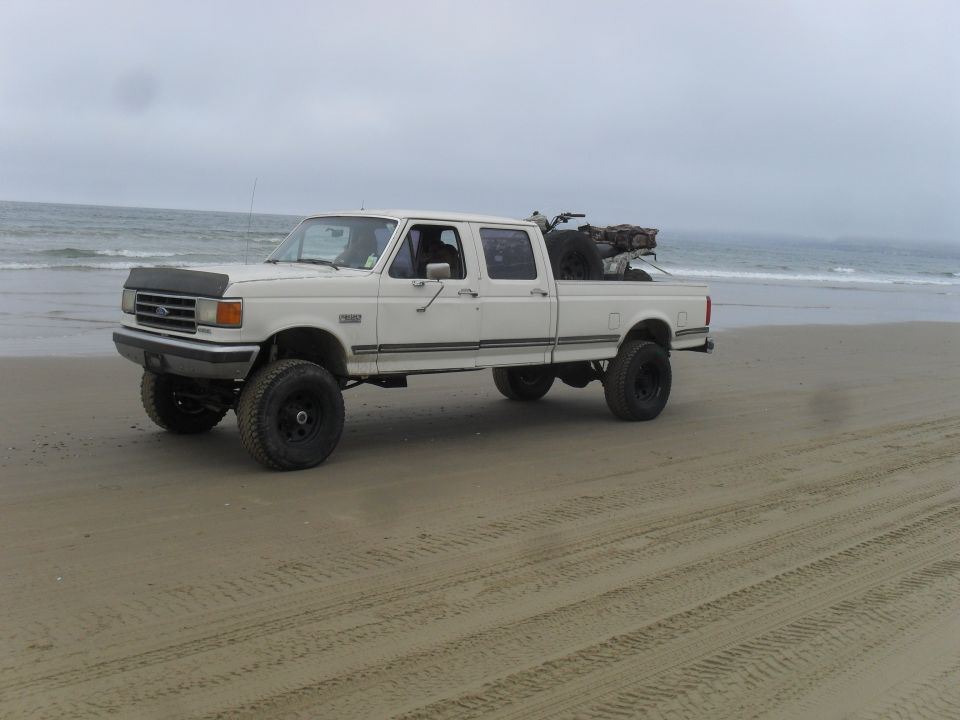 White 1991 Ford F-350 crew cab pickup truck with lifted suspension parked on a sandy beach near the ocean