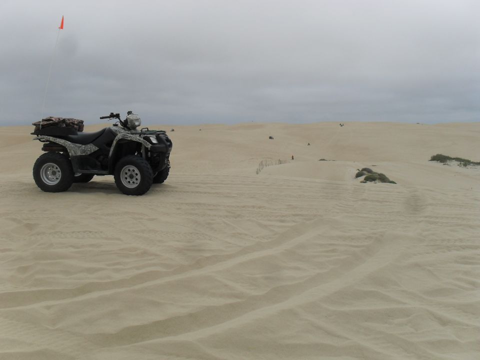 Camouflaged all-terrain vehicle with a storage box on the rear rack and an orange flag on a pole, parked on a sandy dune