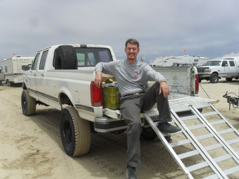 1991 Ford F-350 - A man relaxes in the open tailgate of a white Ford pickup truck, with other vehicles and RVs visible in the sandy background.