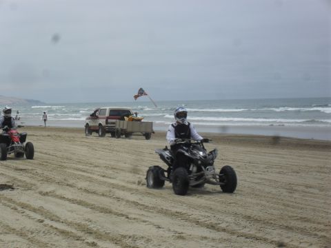 1991 Ford F-350 - ATVs and a pickup truck are driven on a beach with waves in the background.