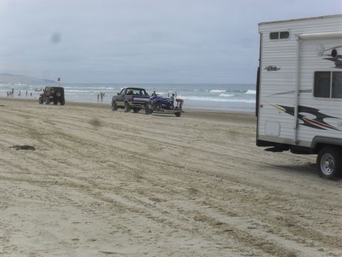 1991 Ford F-350 - A pickup truck tows an ATV on a trailer across a sandy beach near the ocean.