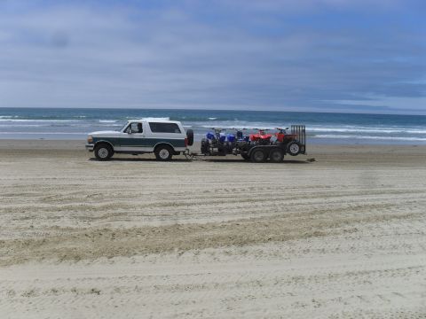 1991 Ford F-350 - A Ford Bronco tows a trailer with ATVs across a beach with the ocean in the background.