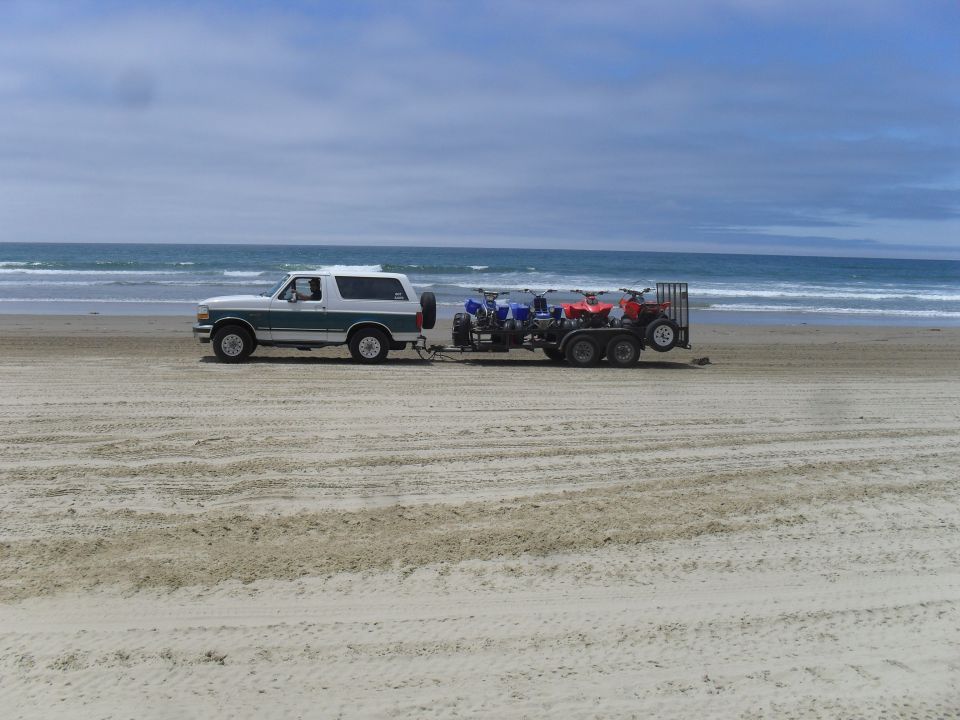 White and green Ford Bronco towing a trailer with ATVs on a sandy beach