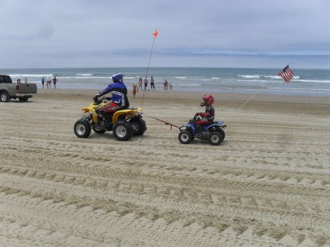 1991 Ford F-350 - Two ATVs are ridden on a sandy beach, with people in the background and an American flag flying.