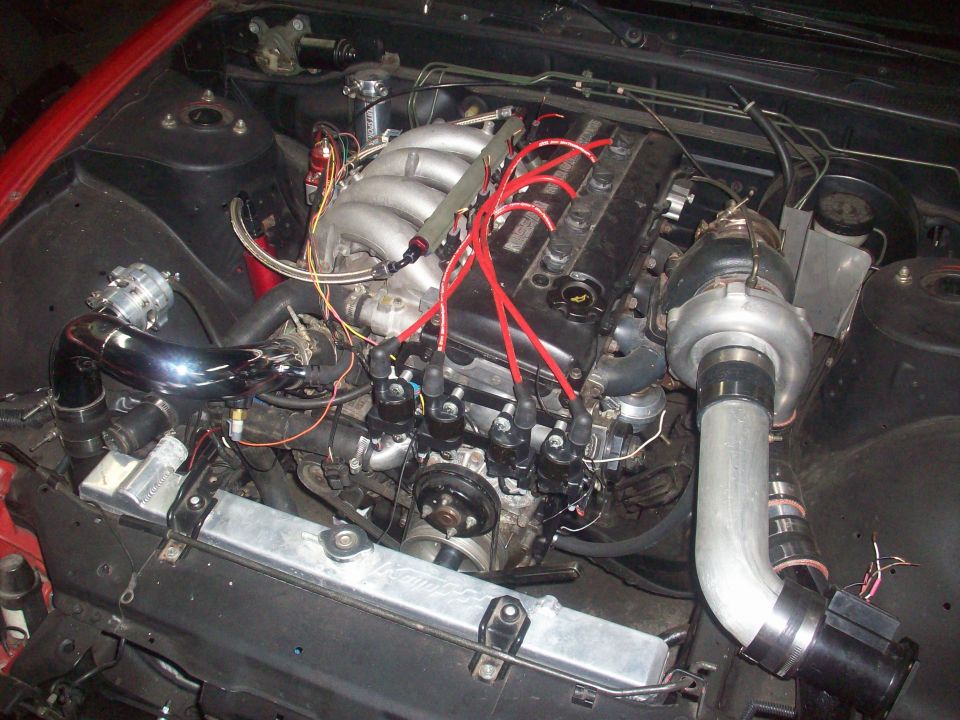 Engine bay with turbocharged inline-six engine, red spark plug wires, silver intercooler piping, and a blow-off valve