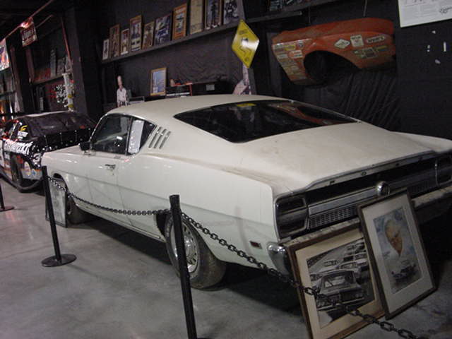 White 1969 Ford Torino coupe, rear quarter panel visible, with a black race car behind it