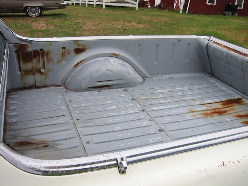 bed of a pickup truck with rust spots and a car in the background