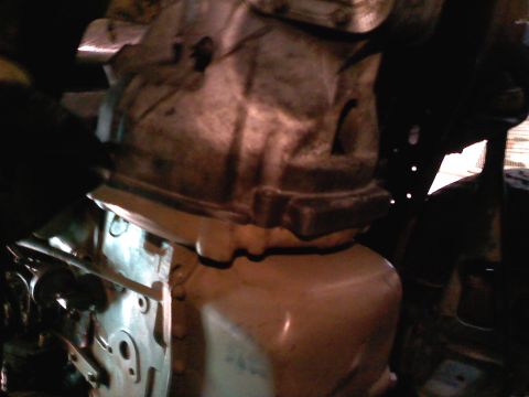 1990 Ford Bronco - Dim close-up looking into an engine bay at the lower rear of a cast-aluminum engine block mated to a tan-painted oil pan, with the bellhous…