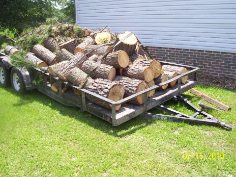 1978 Ford Bronco - A utility trailer is filled with cut logs and branches, parked on a grassy lawn next to a building.