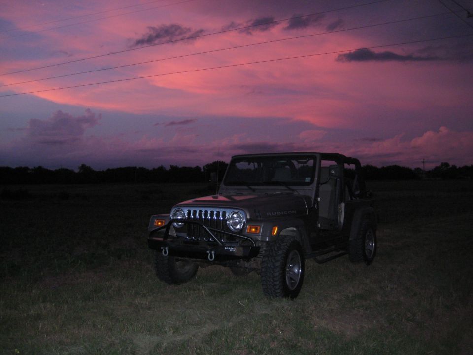 Gray Jeep Wrangler with aftermarket bumper parked in a grassy field under a pink and purple sunset