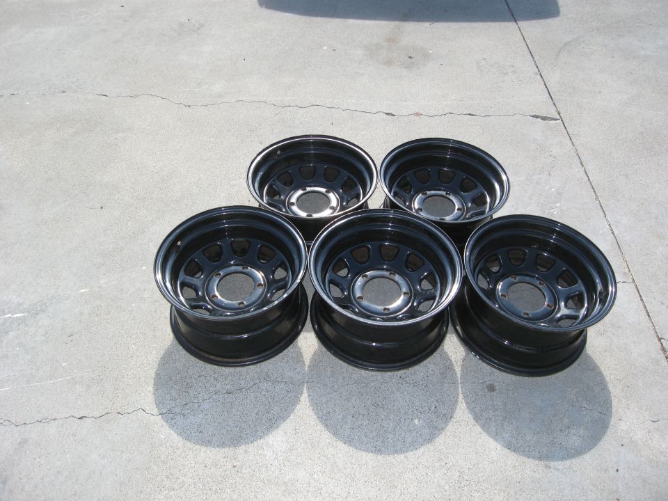 Five black alloy wheels with chrome lips and circular cutouts, arranged on a concrete surface