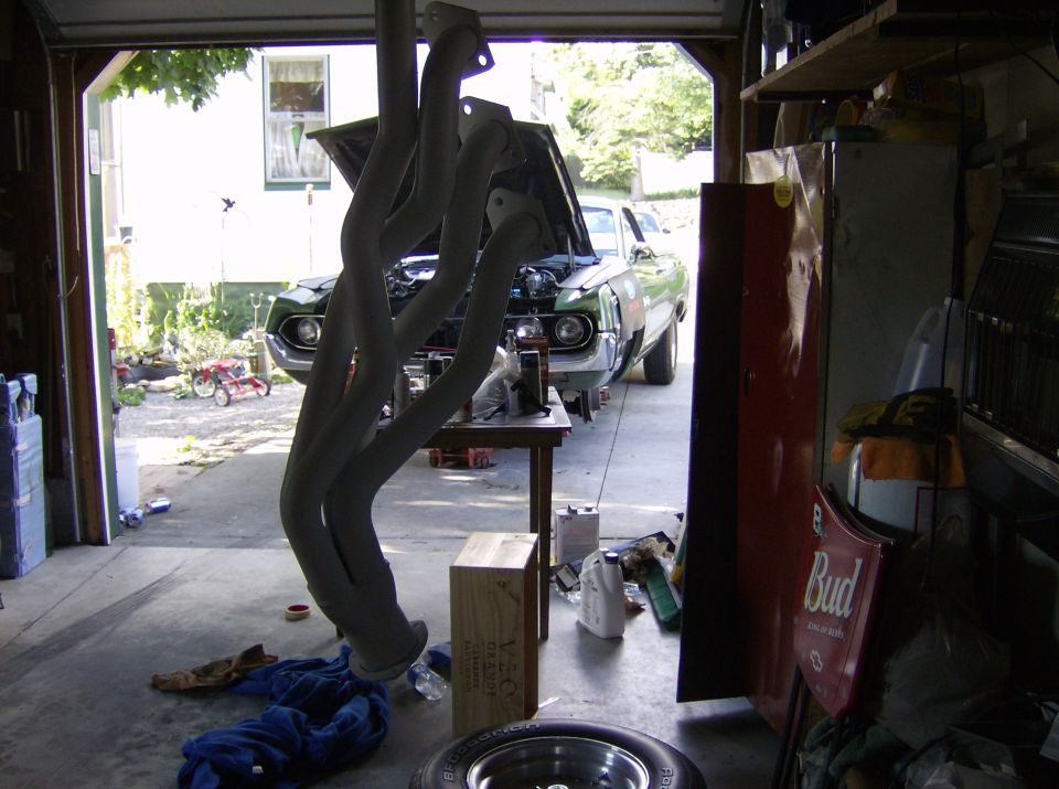 Grey-coated long-tube headers hanging in a garage with a green muscle car behind
