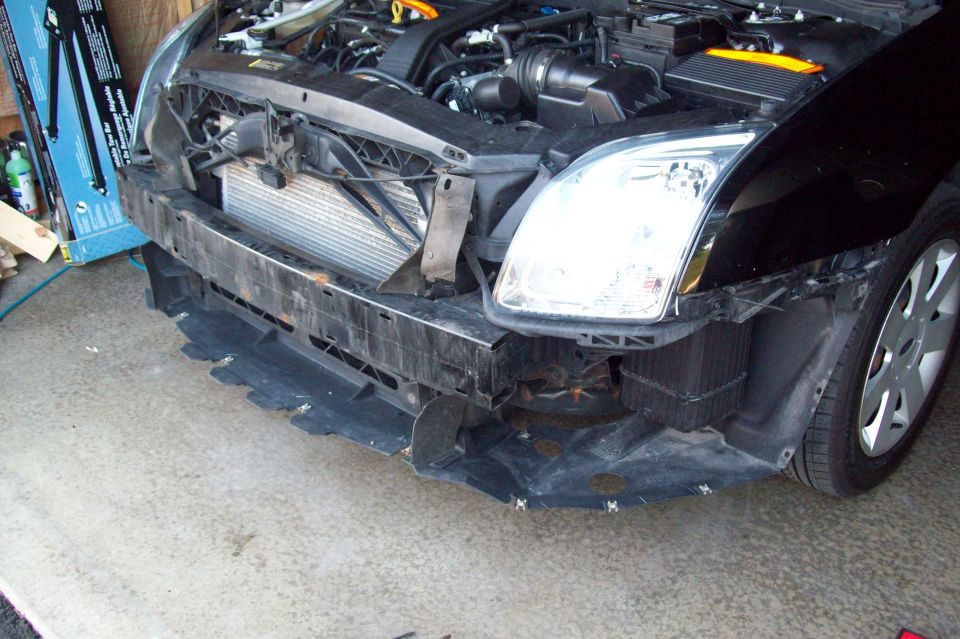 Black car front bumper removed, showing radiator and engine components. Visible wheel and headlight