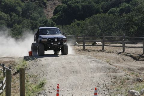 1989 Ford Bronco - A heavily modified off-road truck navigates a dusty dirt track, its large tires creating a cloud of dust as it moves.