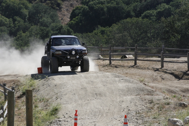 Dark blue off-road truck with large tires and custom bumper driving on a dirt track, kicking up dust