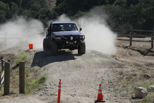 Modified off-road truck driving on a dirt track, kicking up a large cloud of dust. It has large tires and a custom bumper