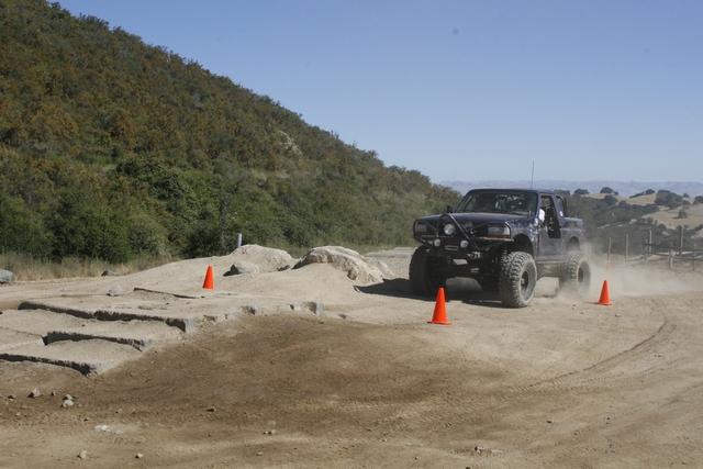 Black off-road truck with large tires driving on a dirt track, kicking up dust