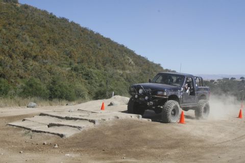 1989 Ford Bronco - Off-road truck navigates a challenging dirt course marked by orange cones, kicking up dust.