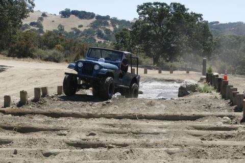 1989 Ford Bronco - A blue off-road jeep tackles a challenging muddy obstacle course on a sunny day.