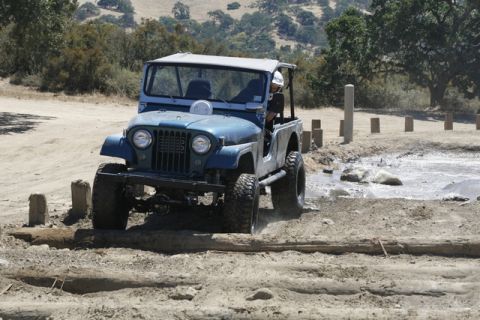 1989 Ford Bronco - Jeep CJ-5 tackles a muddy obstacle course with a driver wearing a helmet.