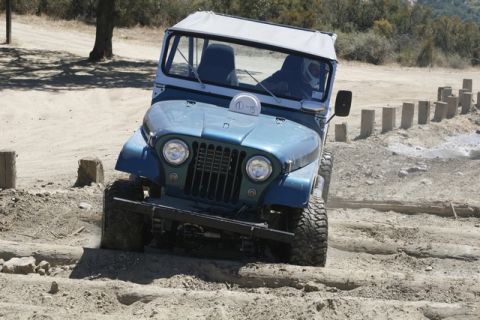 1989 Ford Bronco - A blue Jeep CJ navigates an off-road course with wooden logs and dirt mounds.