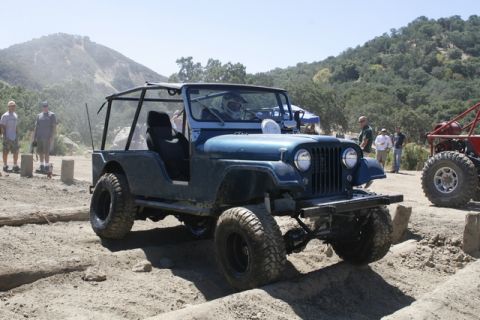 1989 Ford Bronco - A blue Jeep with a roll cage navigates a dirt course, with other vehicles and spectators in the background.