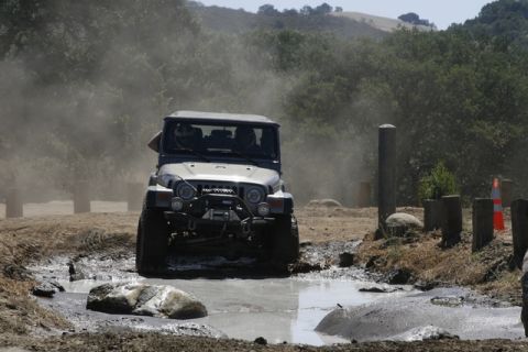 1989 Ford Bronco - Off-road Jeep Wrangler navigates a challenging muddy terrain with dust clouds in the air.