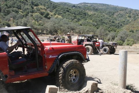 1989 Ford Bronco - Several off-road vehicles, including a red Ford Bronco, are parked on a dirt path with hills in the background.
