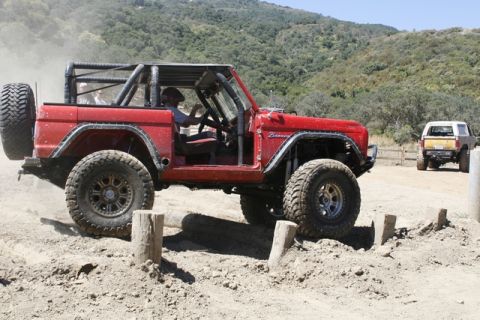 1989 Ford Bronco - A red 1989 Ford Bronco, modified for off-roading with a roll cage, tackles a dirt course with another vehicle in the background.