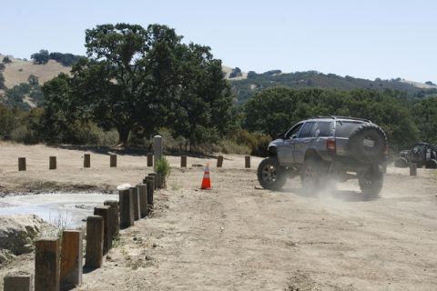 1989 Ford Bronco - SUV drives off-road, kicking up dust on a sunny day with trees and hills in the background.
