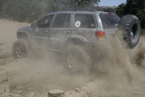 1989 Ford Bronco - A gray Jeep Grand Cherokee navigates a dusty off-road course, its rear tire kicking up a cloud of dirt.