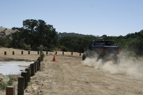1989 Ford Bronco - A dark-colored off-road vehicle drives on a dusty dirt road, creating a cloud of dust behind it.