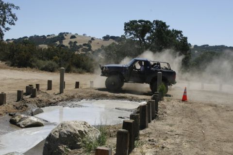 1989 Ford Bronco - Off-road vehicle navigates a muddy obstacle course, creating a cloud of dust.