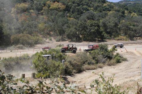 1989 Ford Bronco - Multiple off-road vehicles navigate a dusty dirt track surrounded by trees and hills.