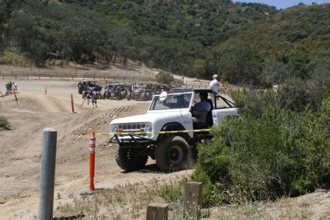 1989 Ford Bronco - A white off-road SUV navigates a dirt track with other vehicles gathered in the distance.