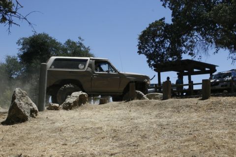 1989 Ford Bronco - A tan SUV navigates rocky terrain under a clear blue sky, with a picnic shelter and another vehicle visible in the distance.