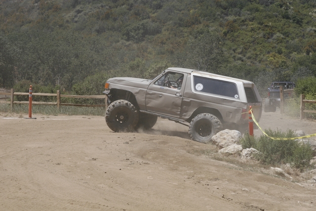 Brown SUV with lifted suspension and large tires driving on a dirt trail, kicking up dust. Another SUV is visible in the background