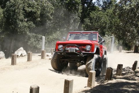 1989 Ford Bronco - A red off-road SUV navigates a dusty dirt path surrounded by trees and wooden posts.
