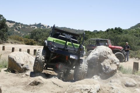 1989 Ford Bronco - Two off-road vehicles tackle a rocky course on a sunny day, with one green vehicle navigating a challenging obstacle.
