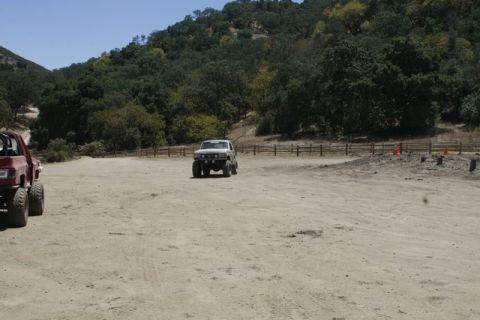 1989 Ford Bronco - Two modified trucks are parked in an outdoor dirt lot with a wooded hillside behind them.