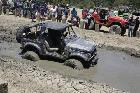 1989 Ford Bronco - Several off-road vehicles are navigating a challenging mud pit with spectators watching.