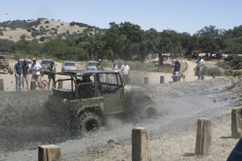 1989 Ford Bronco - An off-road vehicle navigates a muddy track, kicking up spray as onlookers observe the event.