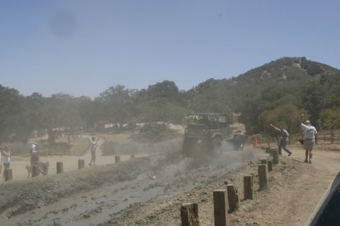 1989 Ford Bronco - A vehicle navigates a muddy track during an event, with onlookers observing from the side.