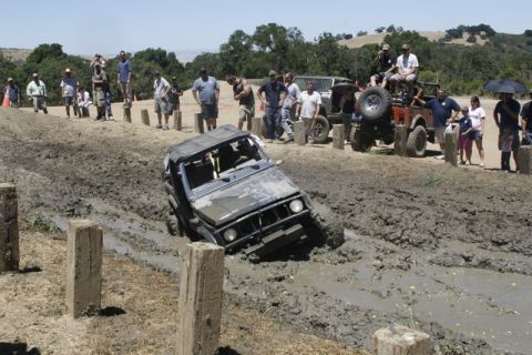 1989 Ford Bronco - An off-road vehicle struggles through a deep mud pit during an event, with onlookers gathered to observe the action.