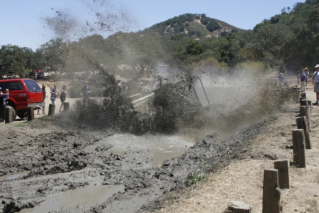 Tube-chassis buggy rolling over in a mud pit, spraying mud as spectators watch