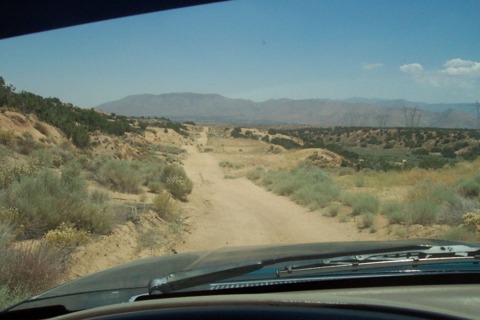 View from inside a vehicle looking out at a dirt road winding through a desert landscape with scrub brush and distant mountains under a clear blue sky