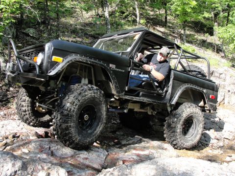 1967 Ford Mustang - A man drives a modified black Ford Bronco over a rocky path in a wooded area.